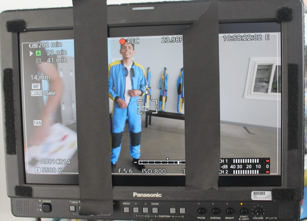 Camera monitor screen showing actor in skydiving uniform and film crew carrying slate